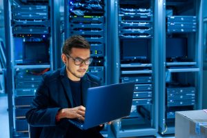 молодой программист в очках за ноутбуком в серверной обновляет ПО Windows VPS